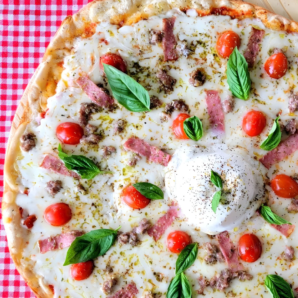 La Piccolina pizza with burrata, cherry tomatoes, basil, sausage, and prosciutto on a red checkered tablecloth.