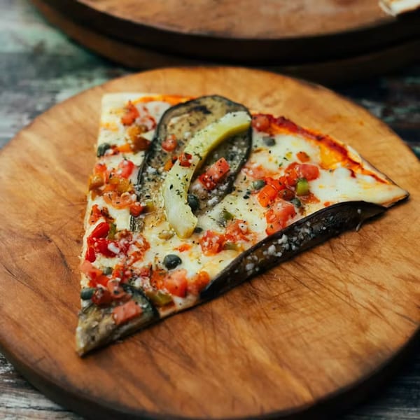 A slice of La Piccolina's pizza with eggplant, tomatoes, and cheese on a wooden board.